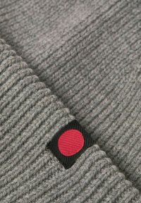 Graue gestrickte Mütze mit geripptem Muster, versehen mit einem schwarzen Etikett, das einen roten, runden Akzent zeigt. Nahaufnahme des Stoffs und der Designdetails.