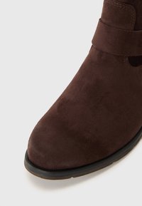 Bottine en daim marron avec un bout rond, dotée d'une sangle décorative et de détails de couture. Texture lisse et subtile brillance à la surface.