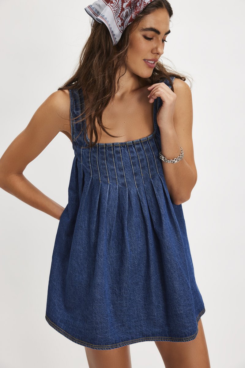 Femme portant une robe en denim bleu sans manches avec des plis, un foulard à motifs et un bracelet chaîne argenté, posant avec une main près du visage.