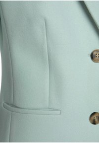 Kabát z vlněné směsi mátově zelené barvy, s klopovým límcem, zapínáním na dva knoflíky a úzkou kapsou na levé straně.