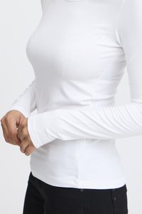 Pitkähihainen valkoinen istuva paita pehmeästä, joustavasta neuloksesta, jossa on pyöreä pääntie ja kapeat hihat, yhdistetty mustiin housuihin.