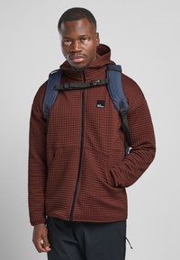 Felpa con cappuccio marrone, trapuntata, con zip e tasche laterali, indossata sopra una maglietta scura, con uno zaino blu e un piccolo logo sul petto.