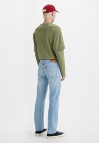 Camisa verde oliva de manga larga recortada, pantalones vaqueros azul claro de pierna recta y zapatillas negras con suelas blancas, vista desde atrás.