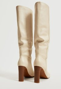 Bottes en cuir beige montant jusqu'au genou, avec une finition lisse, un bout rond et un talon en bois empilé. Détails de couture subtils visibles.