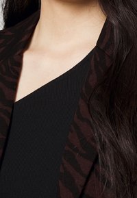 Kaki och svart blusjacka med en texturerad, ribbad halsringning. Har ett mönstrat kavajöverdrag och en vinkelställd krage.