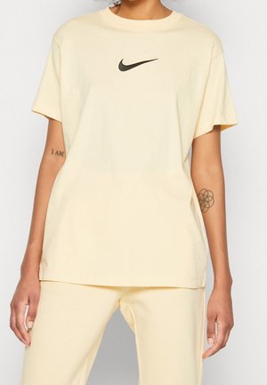 Personne portant un t-shirt Nike couleur crème avec un logo noir centré sur la poitrine et un pantalon assorti, montrant des tatouages sur les deux avant-bras.