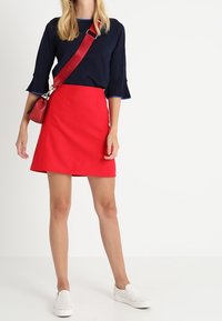 Haut bleu marine à manches longues avec des poignets évasés, associé à une jupe rouge trapèze. Chaussures blanches à enfiler et petit sac bandoulière rouge.