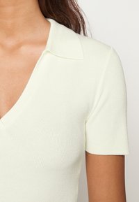 T-shirt de malha canelada amarelo claro, de manga curta, com decote em V e gola clássica. A textura suave acentua o design justo.