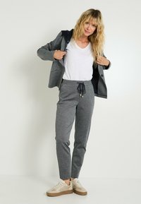 Pantalons de jogging gris en tricot avec une taille à cordon, associés à une veste à capuche grise et noire sur un t-shirt blanc à col en V, et des baskets beiges.