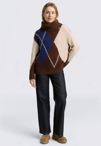 Gebreide trui met een hoge kraag, voorzien van een bruin, crème en blauw ruitmotief. Gecombineerd met donkere spijkerbroek en lichtbruine laarzen.
