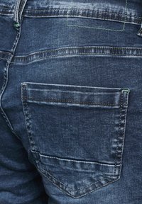 Dark blue denim jeans featuring a back pocket with a reinforced top seam and green accent stitching. Texture is smooth with slight fading.