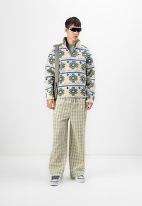 Mies, joka on pukeutunut kuviolliseen fleece-takkiin, leveisiin ruutuhousuihin, lenkkareihin, aurinkolaseihin ja olkalaukkuun, vaaleaa taustaa vasten.