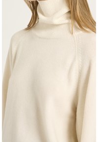 Kobieta ubrana w kremowy sweter z golfem o prążkowanej fakturze, z długim rękawem, pokazana od szyi do połowy tułowia.