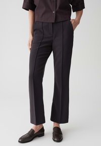 Pantalons de costume sombres avec une coupe ajustée, présentant un devant plissé, des jambes droites et une texture lisse. Portés avec des mocassins marron et une chemise à col.