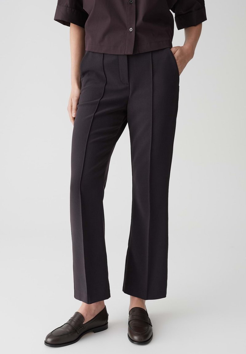 Pantalons de costume sombres avec une coupe ajustée, présentant un devant plissé, des jambes droites et une texture lisse. Portés avec des mocassins marron et une chemise à col.