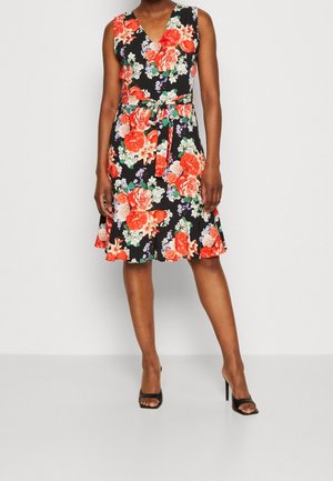 Frau trägt ein ärmelloses schwarzes Kleid mit rotem und weißem Blumenmuster, gebundenem Taillengürtel und schwarzen offenen High Heels.