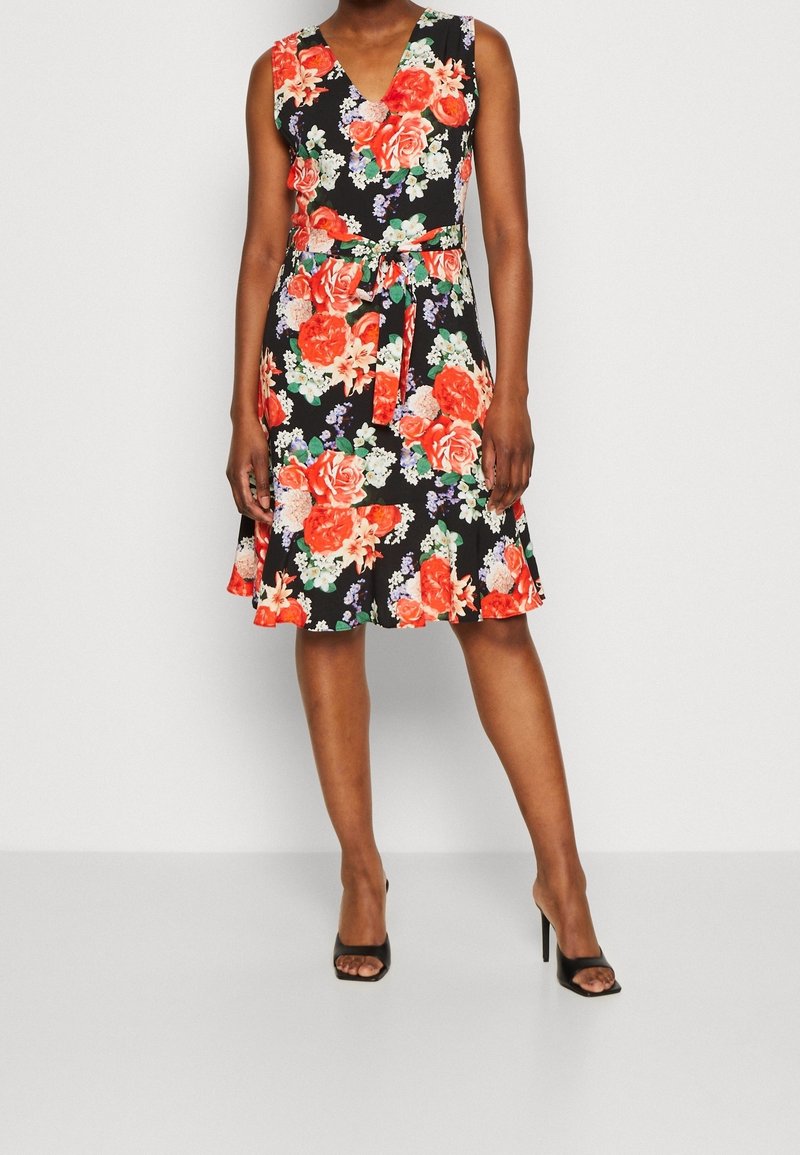 Femme portant une robe noire sans manches à imprimé floral rouge et blanc, ceinture nouée à la taille et escarpins noirs à bout ouvert.