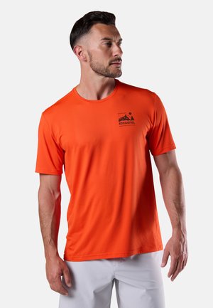 Uomo con capelli corti e scuri che indossa una maglietta arancione Rossignol e pantaloni grigio chiaro, che guarda verso sinistra su uno sfondo semplice.