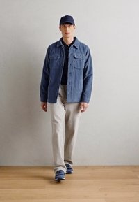Casaco de veludo cotelê azul com dois bolsos no peito sobre uma camisa preta, combinado com calças bege claras e ténis azuis, em frente a uma parede neutra.