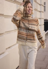 Pull en maille à col montant, présentant un motif marron et crème, un design textur�é et une bordure côtelée. Associé à un pantalon de couleur claire.