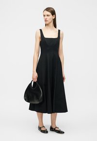 Femeie purtând o rochie midi fără mâneci, neagră, cu decolteu pătrat, ținând o geantă rotundă neagră și purtând pantofi negri Mary Jane.