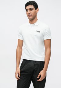 Fehér póló klasszikus gallérral és rövid ujjakkal, a mellkasán fekete "KARL LAGERFELD" logóval. Fekete nadrággal viselve.