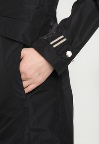 Manica di giacca nera con chiusura a bottoni e dettagli a righe beige. Il tessuto è liscio con una leggera lucentezza e un design lineare.