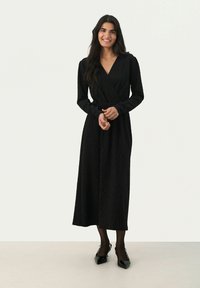 Schwarzes Wickelkleid mit langen Ärmeln, strukturiertem Stoff, der ein Zickzackmuster aufweist, und einer taillierten Passform, kombiniert mit glänzenden schwarzen High Heels.