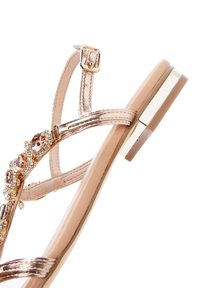 Sandalias con tiras en un metálico dorado rosa, adornadas con piedras preciosas y con un tacón cuadrado. Presentan una correa ajustable en el tobillo y una suela beige.