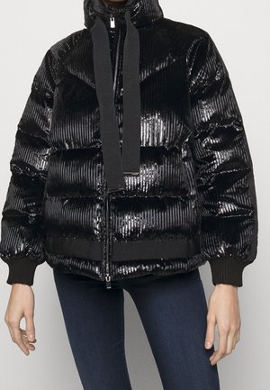 Kobieta w błyszczącej, czarnej, prążkowanej kurtce puchowej z kapturem, dużymi ściągaczami i dopasowanych ciemnoniebieskich spodniach.