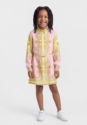 Usmievavá dievčina s vrkočmi, oblečená v ružových a žltých šatách s opaskom a bielymi teniskami, stojaca pred jednoliatym pozadím.