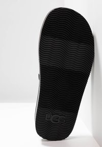 Semelle d'une chaussure UGG noire avec un motif ondulé, présentée sur fond blanc, mettant en avant l'adhérence texturée et le logo de la marque.