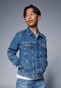 Denim jakna v srednje modri barvi, z značilnim dizajnom, ki vključuje dva žepa na prsih, zapiranje z gumbi in srednje oprano teksturo.