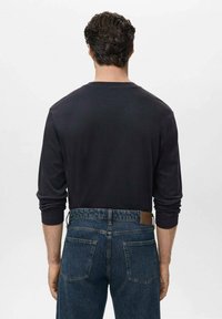 Dunkles Langarmhemd, kombiniert mit blauen Jeans. Das Hemd hat einen Rundhalsausschnitt und einen lockeren Schnitt; die Jeans verfügen über Gesäßtaschen und ein braunes Lederetikett.