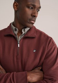 Tmavorudá mikina na zips so stojatým golierom, rebrovanými manžetami a malým vyšívaným logom vtáka na hrudi. Hladká textúra látky.