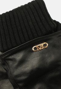 Cuello de chaqueta de cuero negro con parte superior de punto negro acanalado y un detalle de logo en tono dorado. Superficie texturizada con costuras visibles.