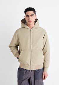 Sweat à capuche beige zippé avec une capuche doublée, des poches avant et des poignets côtelés. Associé à un pantalon cargo gris avec des panneaux texturés.