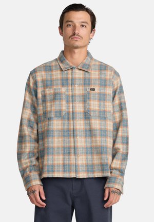 Homme portant une chemise à carreaux beige, bleue et orange avec deux poches poitrine et un pantalon foncé, sur un fond uni.