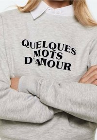 Felpa grigia con la scritta "QUELQUES MOTS D'AMOUR" in carattere nero in grassetto. Dettagli comprendono polsini a costine e scollo rotondo.