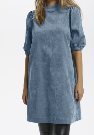 Robe en velours côtelé bleu avec manches courtes bouffantes, col montant et coupe ample. La texture est douce avec un léger côtelage, se terminant juste au-dessus des genoux.