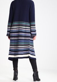 Długi granatowy kardigan z pionowymi paskami w zielonym, niebieskim i różowym kolorze. Dzianinowa faktura, dekolt w serek i długie rękawy. Sięga poniżej kolan. Czarne botki za kostkę.