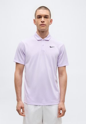 Jeune homme aux cheveux courts portant un polo Nike violet clair et un short blanc, debout devant un fond blanc uni.