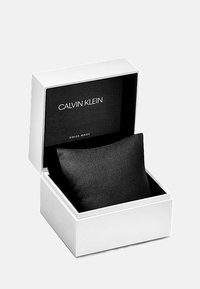 Boîte de montre Calvin Klein en blanc avec un intérieur noir, contenant un coussin noir rembourré. Présente le texte : "CALVIN KLEIN" et "SWISS MADE."