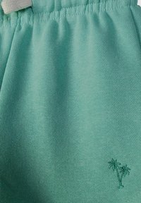 Gros plan sur un short en tissu bleu sarcelle avec une taille élastique et deux petits palmiers brodés près du bord inférieur.