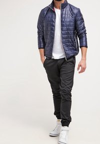 Giacca imbottita blu navy con colletto alto, zip frontale e cuciture orizzontali, abbinata a pantaloni neri a gamba stretta e sneakers bianche alte.