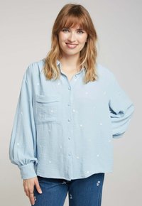 Chemise bleu clair à boutons, en tissu texturé, avec une poche plaquée et un broderie florale blanche sur toute la surface. Manches longues.