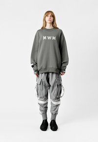 Sudadera gris de gran tamaño con la letra "MWM", combinada con pantalones cargo grises con cremalleras y acentos blancos. Calzado negro texturizado.