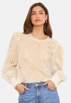 Kobieta ubrana w beżową bluzkę z długim rękawem z kwiatowymi wzorami wycinanymi i falistymi krawędziami, w połączeniu z niebieskimi jeansami, ręce w kieszeniach.