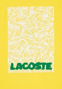 T-shirt jaune avec un carré blanc affichant un motif répétitif de crocodiles et le texte "LACOSTE" en gras vert en bas.
