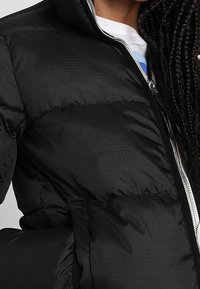 Personne portant une veste matelassée noire à moitié zippée par-dessus une chemise blanche, avec des cheveux tressés visibles sur le côté droit.